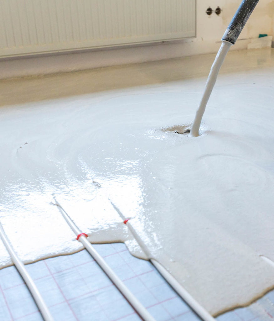 Workman pouring pumped liquid floor screed in a residential property, covering underfloor heating pipes and insulation panels